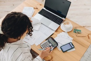 view of a woman working at home in the kitchen with financial papers, counting on a calculator, paying bills, planning a budget to save some money