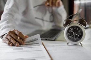 selective focus on alarm clock, asian businessman review business document using laptop working on table before work time end in a day